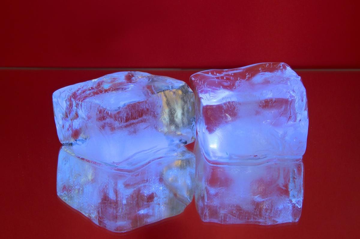 Two ice cubes sit on a reflective surface in front of a red background