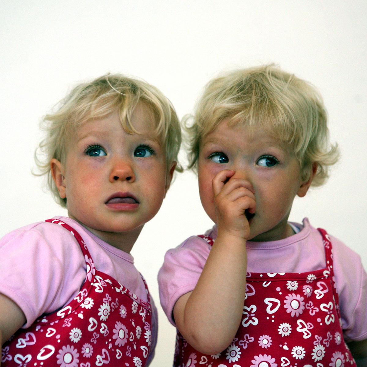 A pair of blonde twin toddlers look in opposite directions from the camera