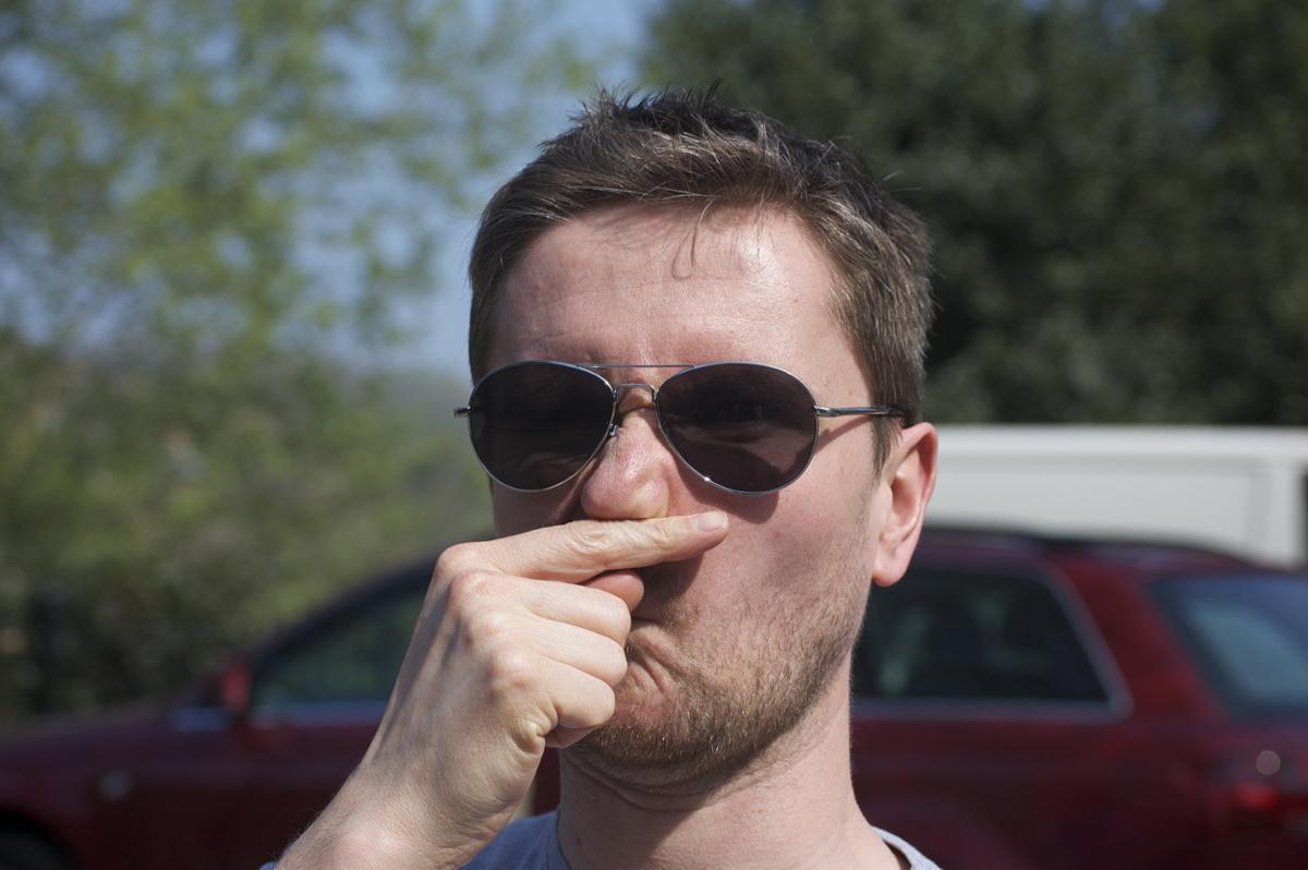 A man standing outside with sunglasses on rubs his finger under his nose to scratch it