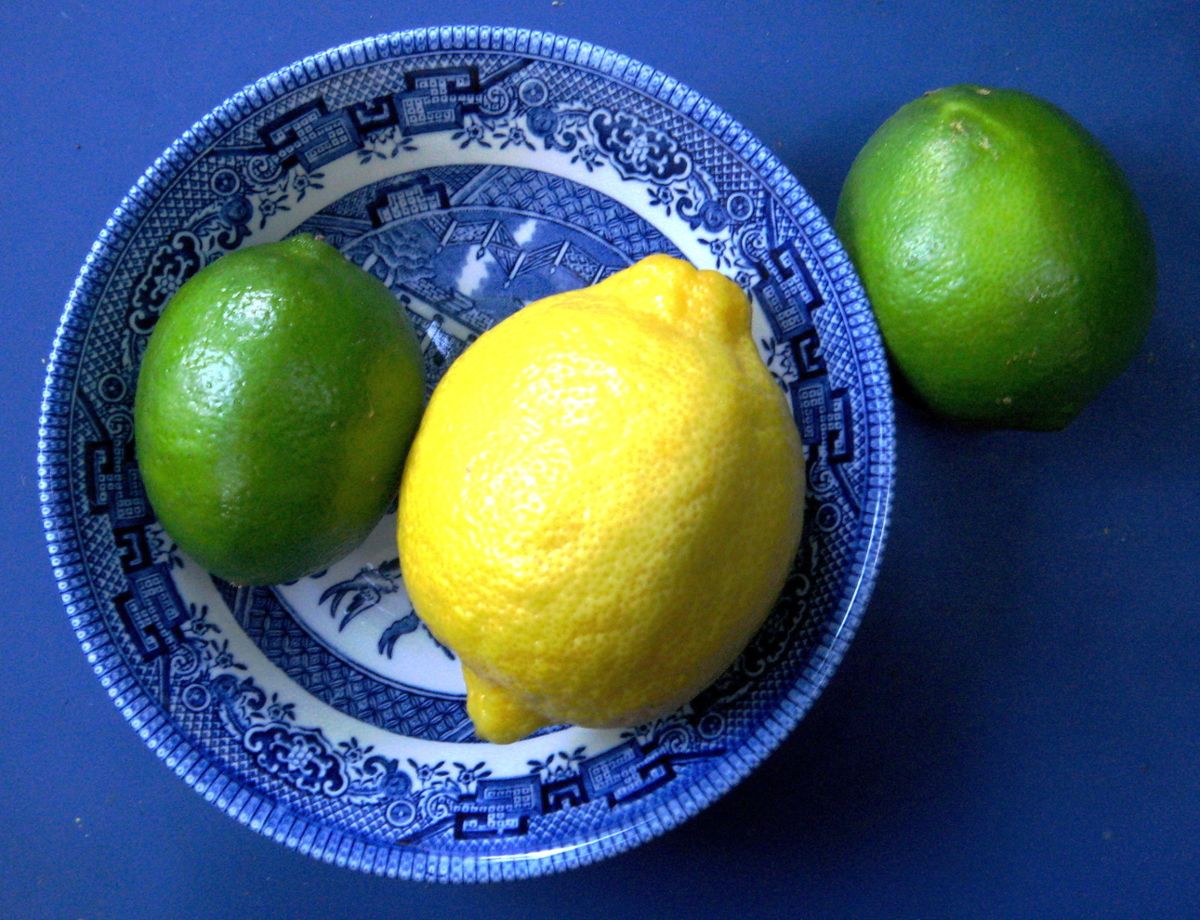 A lemon and a lime inside a blue bowl on a blue surface, with another lime outside of the bowl