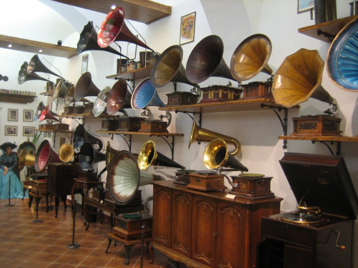 A collection of a variety of phonographs sit displayed against a white wall