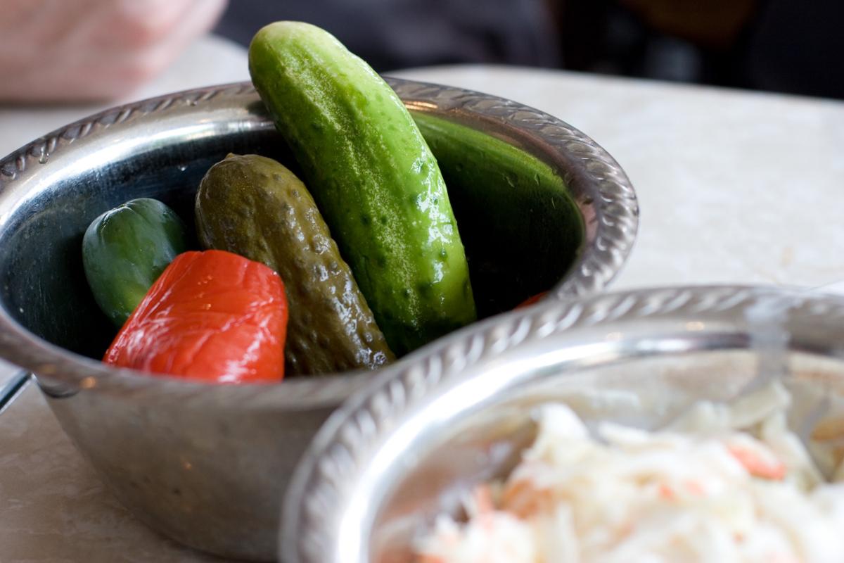 Pickles and a red pepper sit in a silver bowl as part of a larger meal
