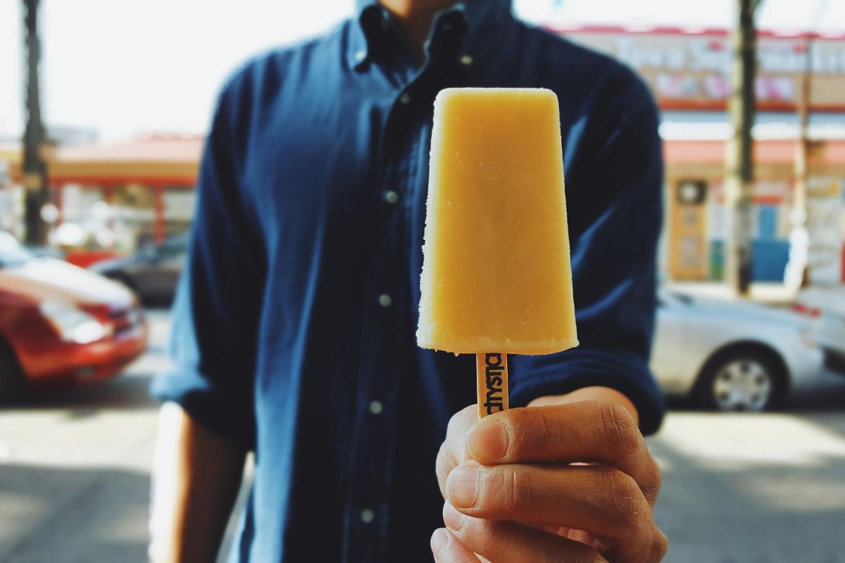A man in a blue shirt offers a yellow popsicle to the camera while standing on the sidewalk of a street