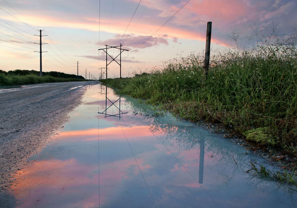 A large puddle on the side of the road, with a pink and blue sky over power lines