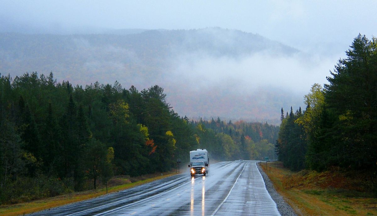 A truck pulling a camper down a rainy road in a wooded area, with a foggy mountain behind