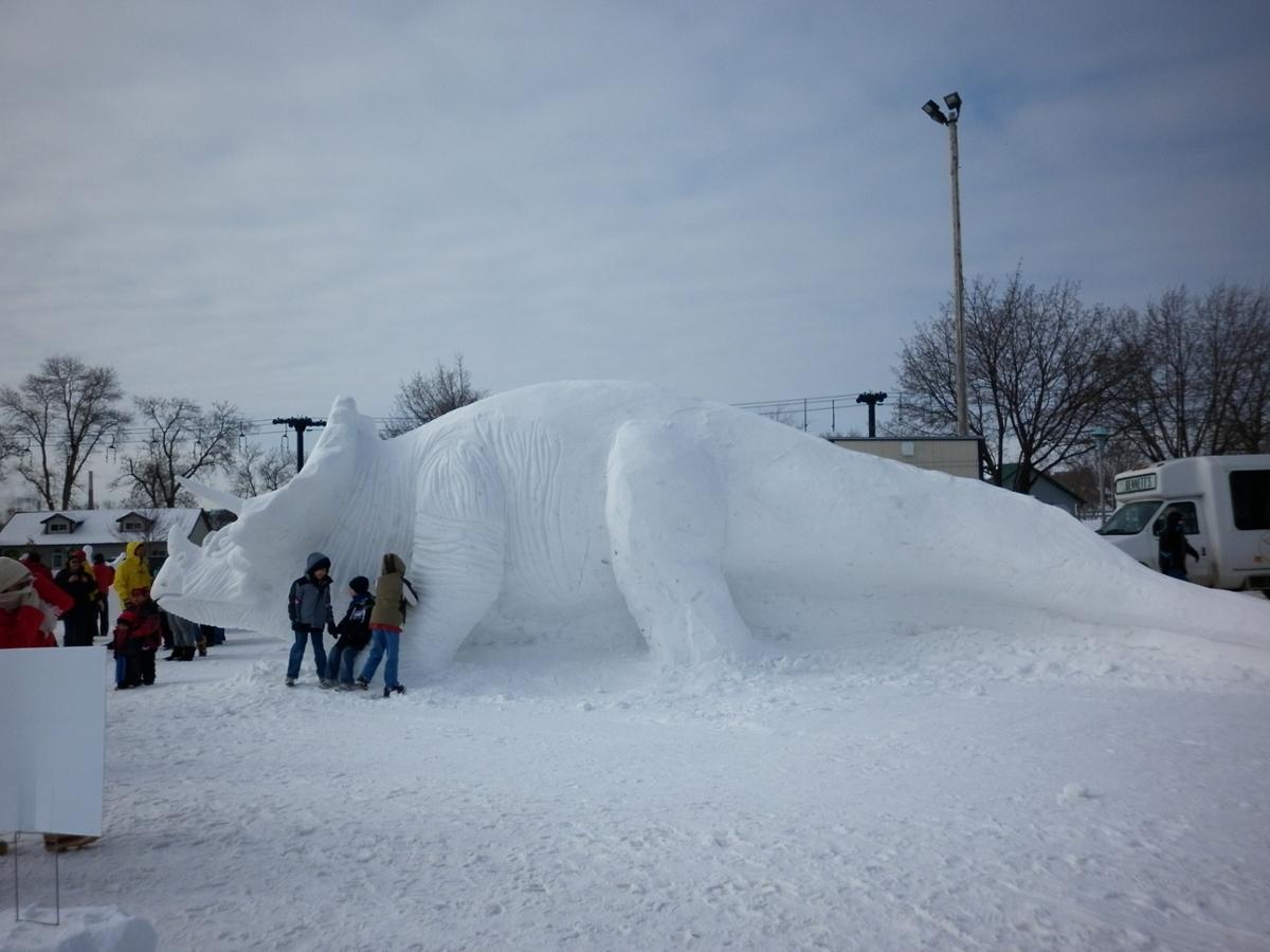 A triceratops sculpture made out of snow, with a small group of children standing next to the head for scale