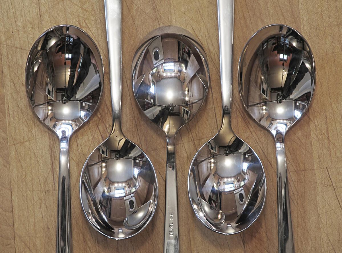 Five spoons are arranged on a wooden surface with a kitchen reflected in their surfaces