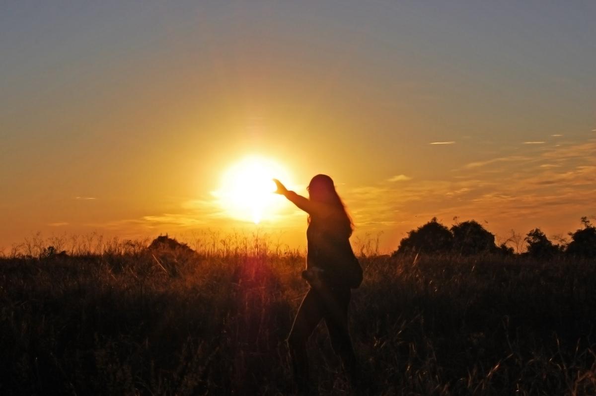 A woman stands in silhouette in front of the setting sun with her hand outstretched 