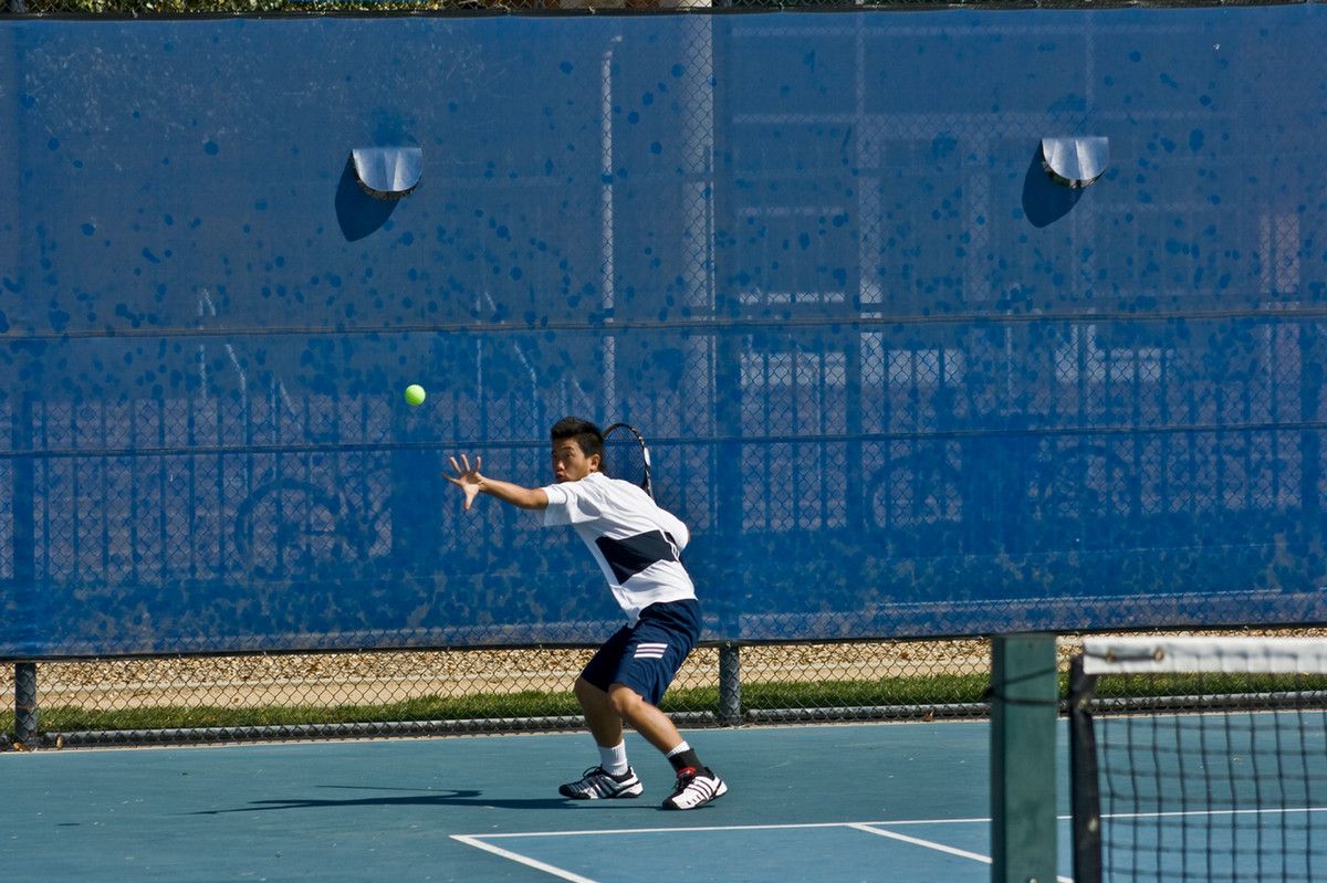 A man playing tennis against a blue backdrop, the ball up in the air and his racket raised to hit it