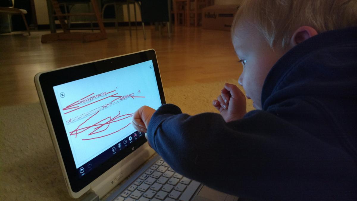 A toddler lays on the floor with a touch screen tablet in front of them, drawing random lines