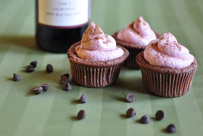 Wine-flavored cupcakes with pink frosting sit next to a red wine bottle and scattered chocolate chips