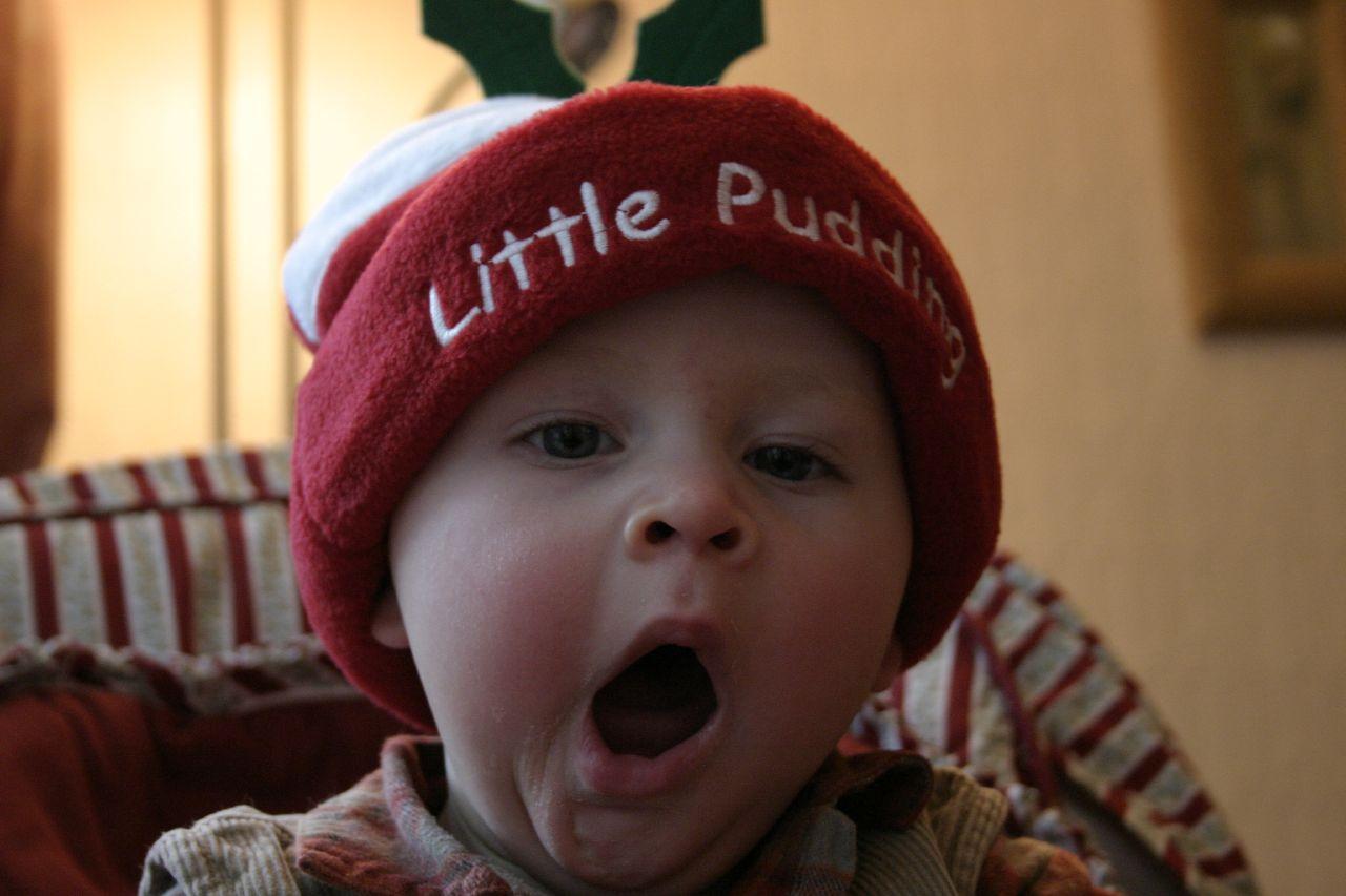 A baby with a large red hat that reads "little pudding" yawns while looking at the camera
