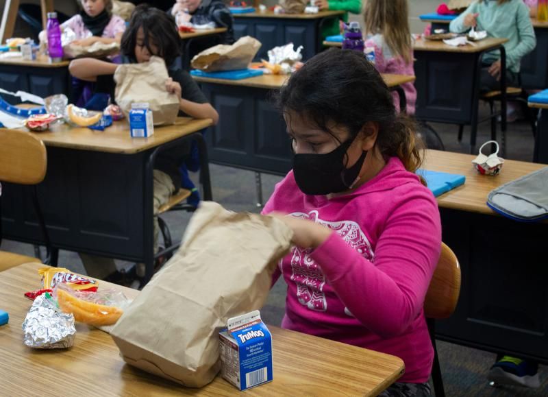 A girl in a bright pink shirt with a black face mask unpacks a sack lunch at a desk in a classroom. Other students are in the background