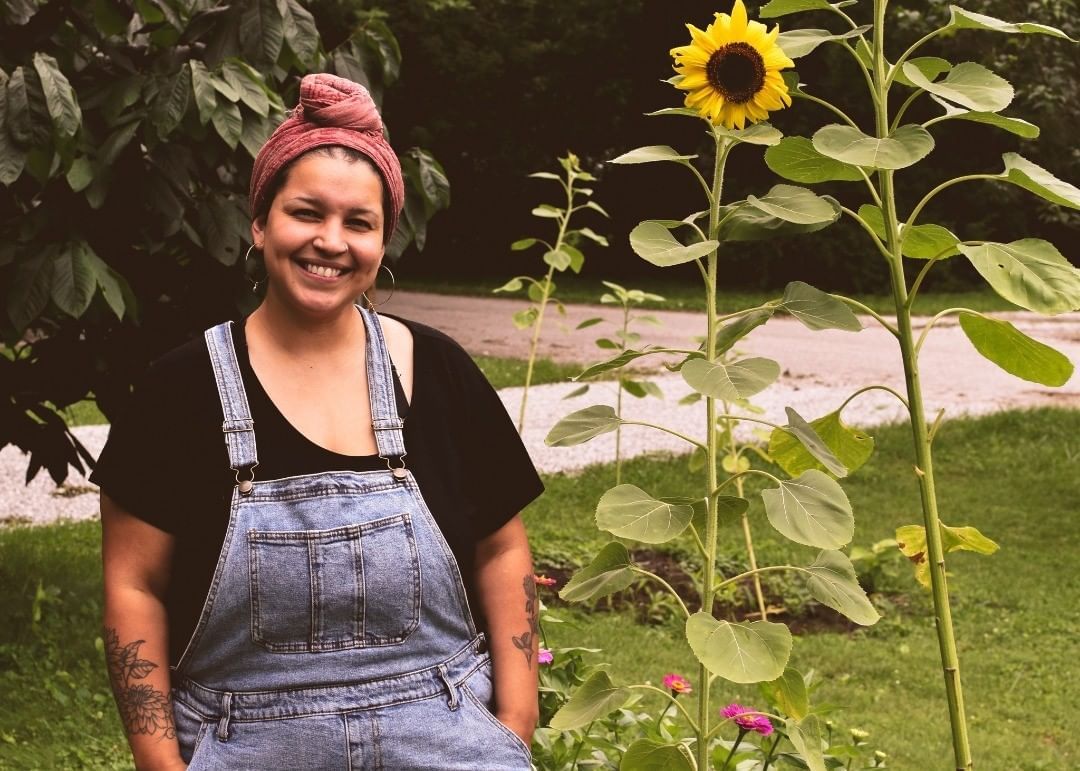 Shanna Hughey in denim overalls with hair bound in a scarf standing next to a tree and sunflower, smiling at camera