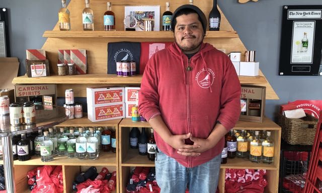 Abel Garcia in a red sweatshirt standing in front of a wooden display of colorful bottles of spirits