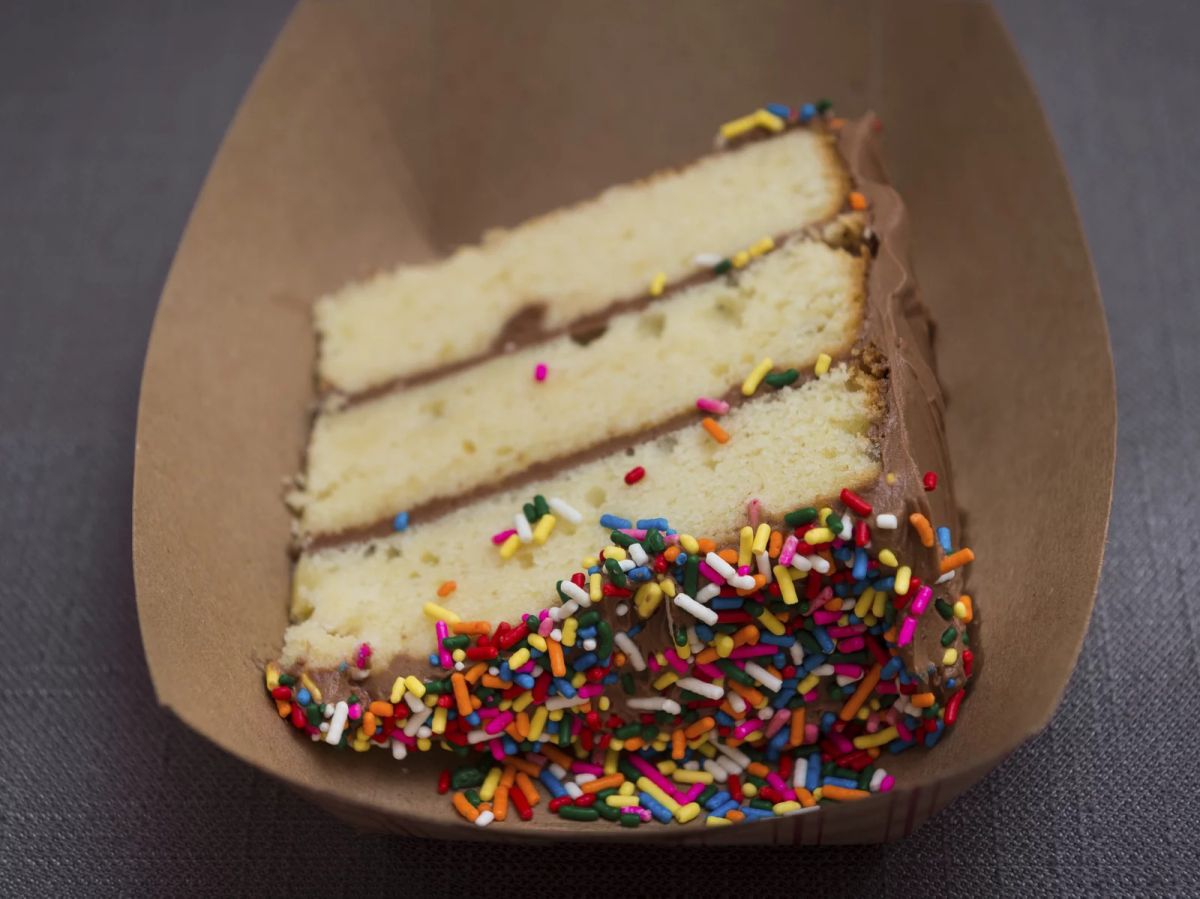 A piece of vanilla cake with chocolate frosting and multicolor sprinkles in a brown cardboard serving tray