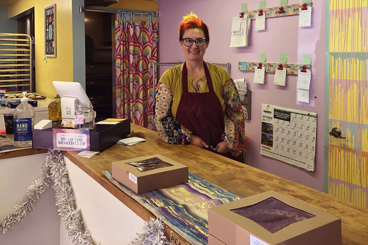 Brittany Kiel, with brightly colored hair, standing at a long counter, with pink and yellow walls, a brightly patterned curtain in the background. 