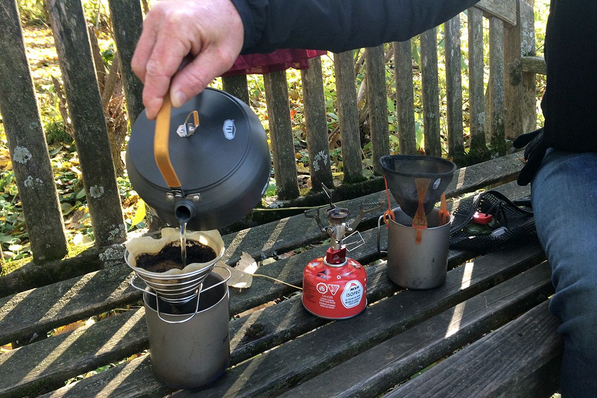 A hand pouring a small kettle of water over a pour-over device for coffee on a wooden bench outdoors.
