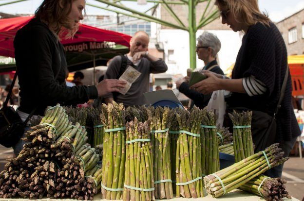 A woman handing money to another woman at a farm stand with asparagus in view