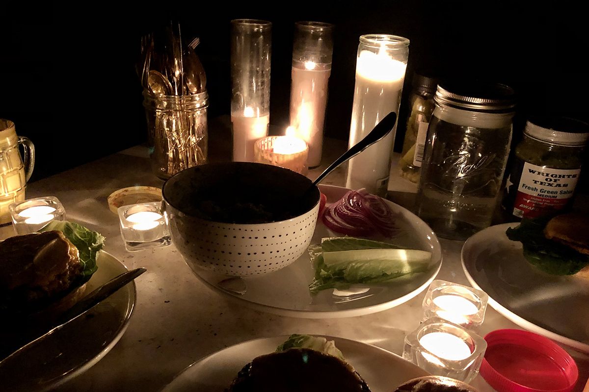 A table top, in the dark, with dishes and several white votive candles, silverware in a mason jar, lettuce and onion visible on a plate