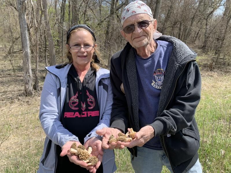 A white middle aged woman and a white elderly man staning outside looking at the camera with wrinkly mushrooms in their hands and woods behind them