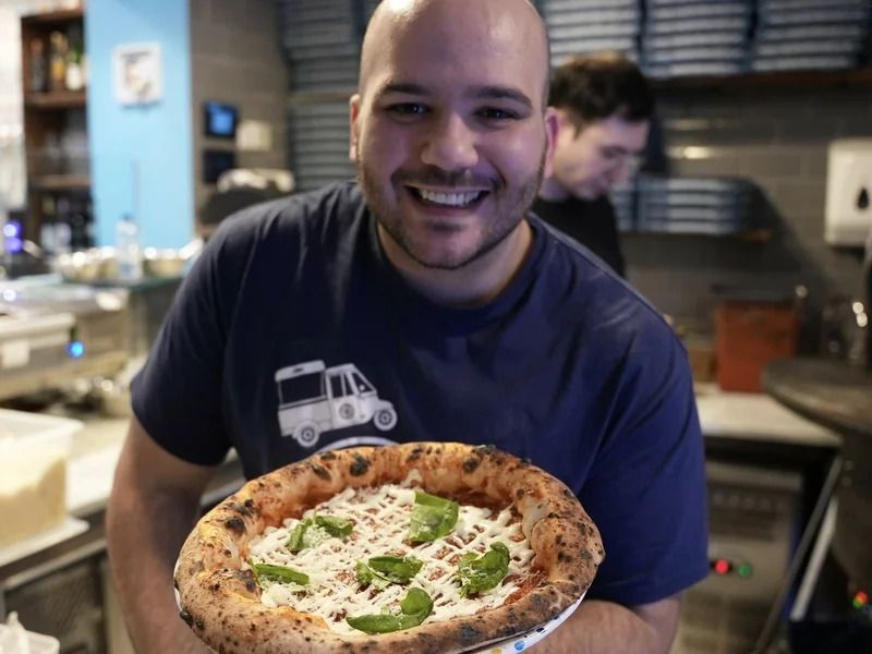 Michele Pascarella holding pizza smiling at camera inside restaurant setting