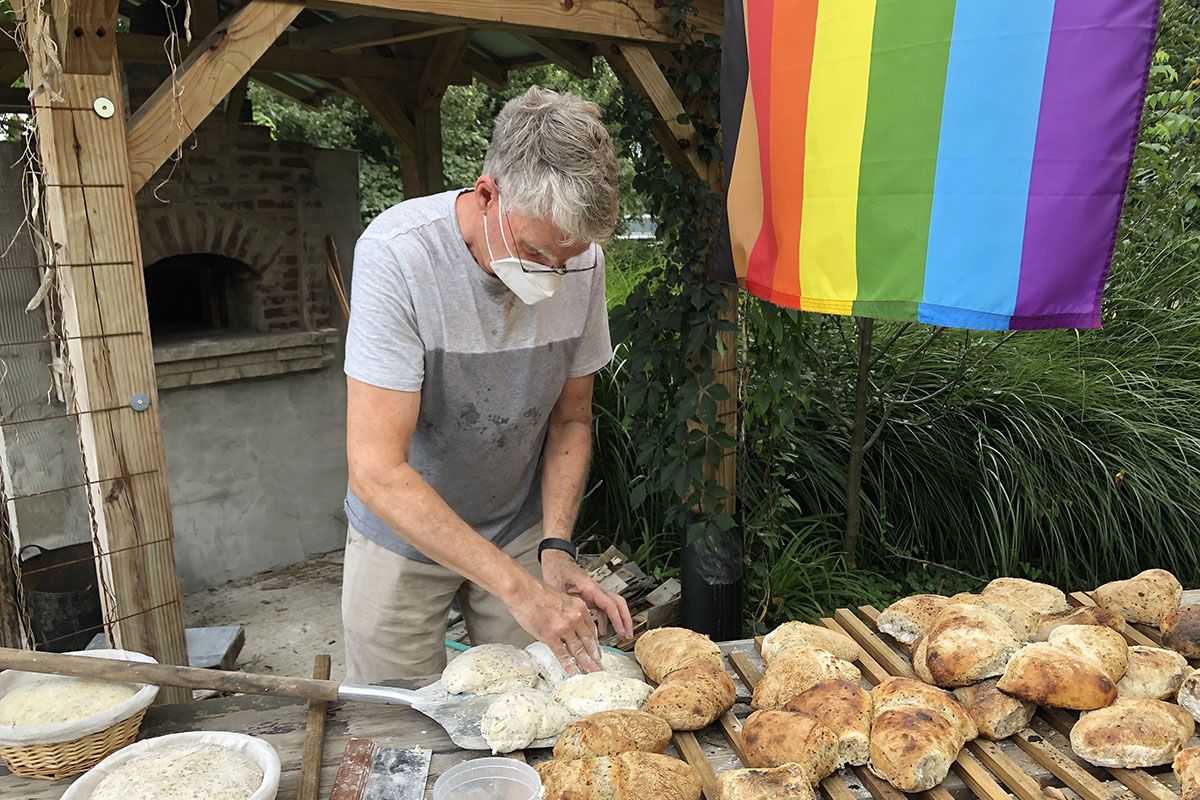 Keith Romaine wearing face mask cutting bread dough with baked loaves and baskets of dough on table, outdoor brick oven behind him and rainbow flag above.