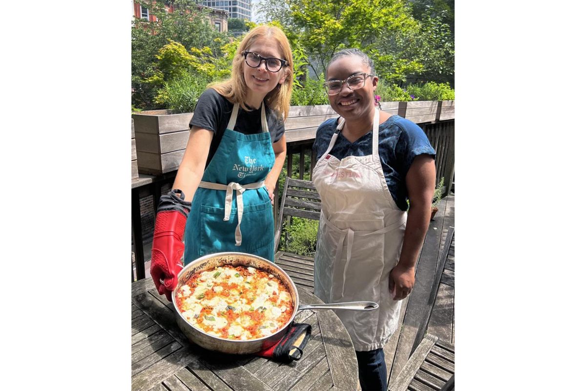 Melissa Clark and Ayesha Rascoe in aprons with a dish of food on a patio outside