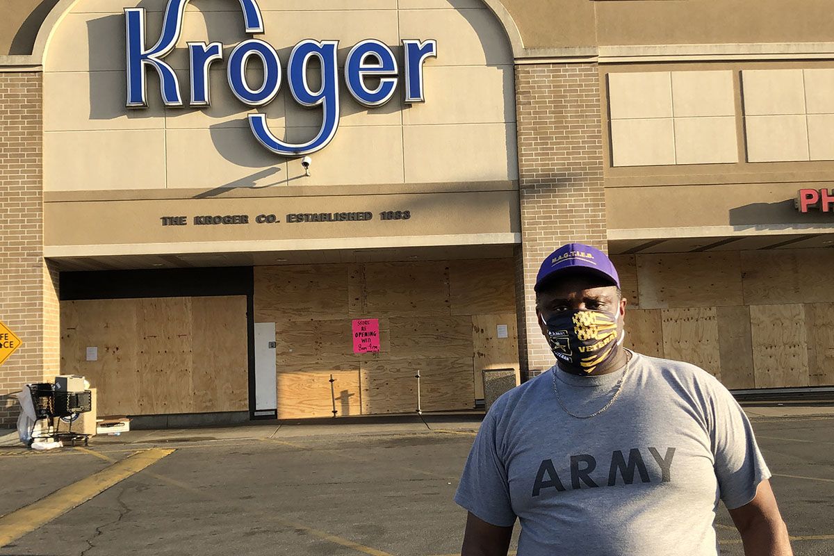 MeShorn Daniels with face mask and Army shirt, in ball cap looking at camera standing in parking lot of boarded-up Kroger.