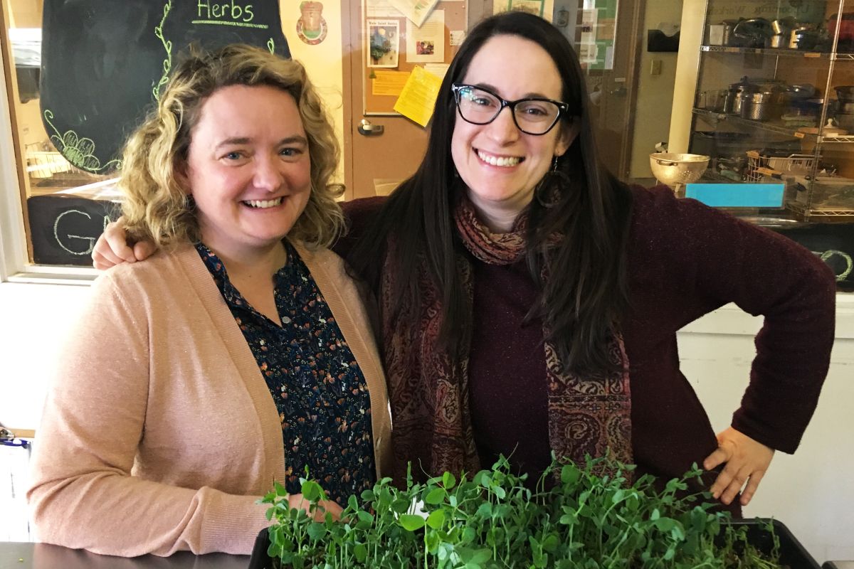 Amanda Nickey and Stephanie Solomon smiling at the camera with a tray of micro-greens in front of them and a window onto a kitchen behind them. 