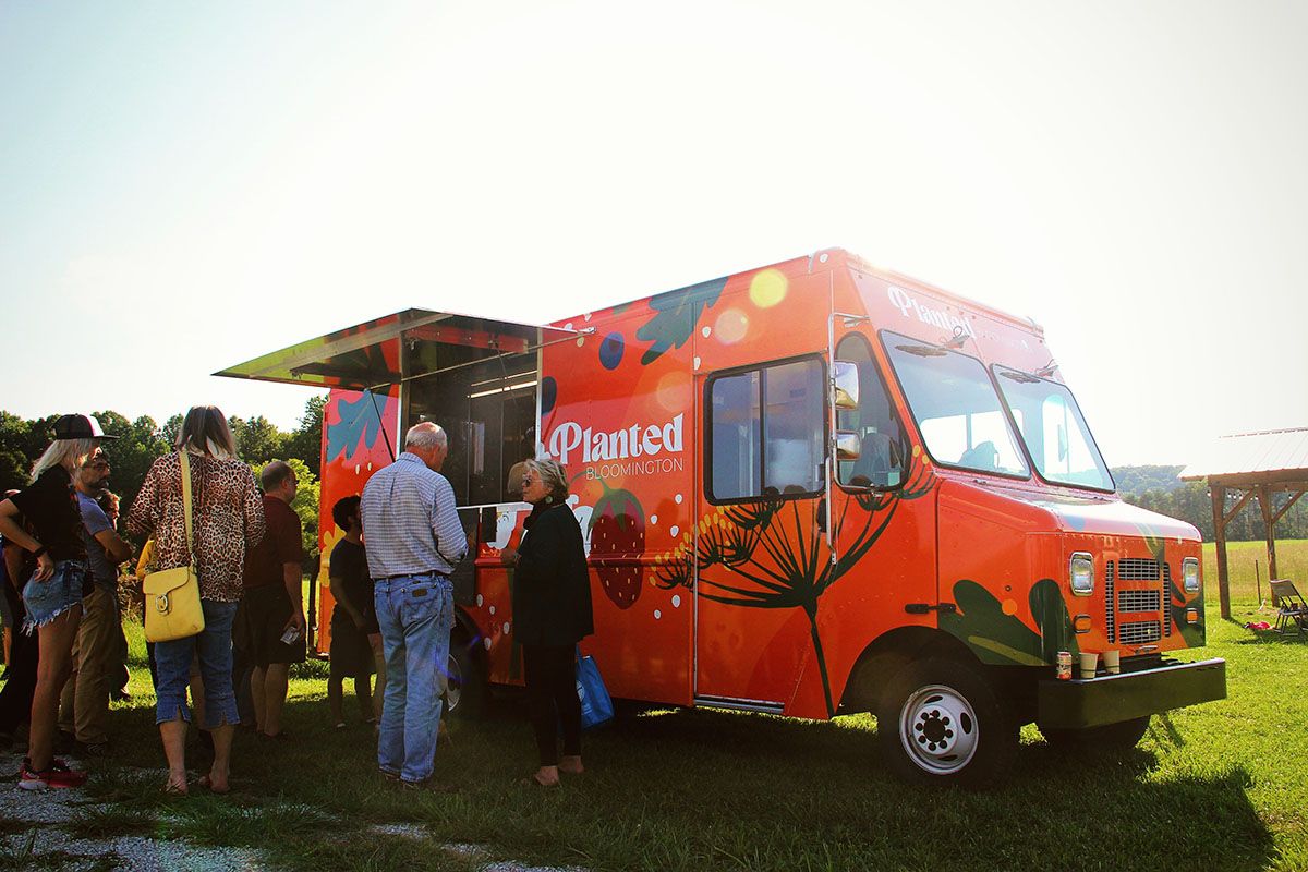 people lined up at an orange food truck. the truck has large, colorful graphics of plants like dill flowers, chard leaves, strawberries, etc.