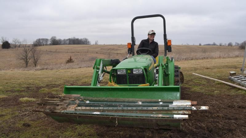 Dusty Spurgeon, facing the camera, smiling, driving a green tractor with front-loader filled with fence posts. 