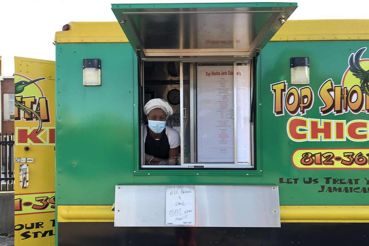 Taneisha Henline with face mask and scarf around hair, in window of yellow and green food truck