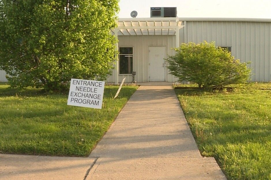 An entrance to a needle exchange program.  A small, simple yard sign points to the entrance past a sidewalk and tan building.