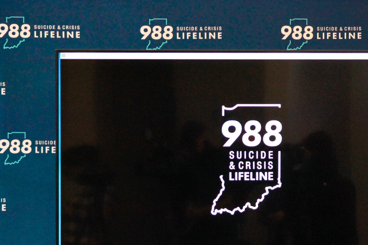 A large black screen has the 988 hotline logo in its center. Behind it is a blue backdrop with the 988 hotline logo stamped across it repeatedly.