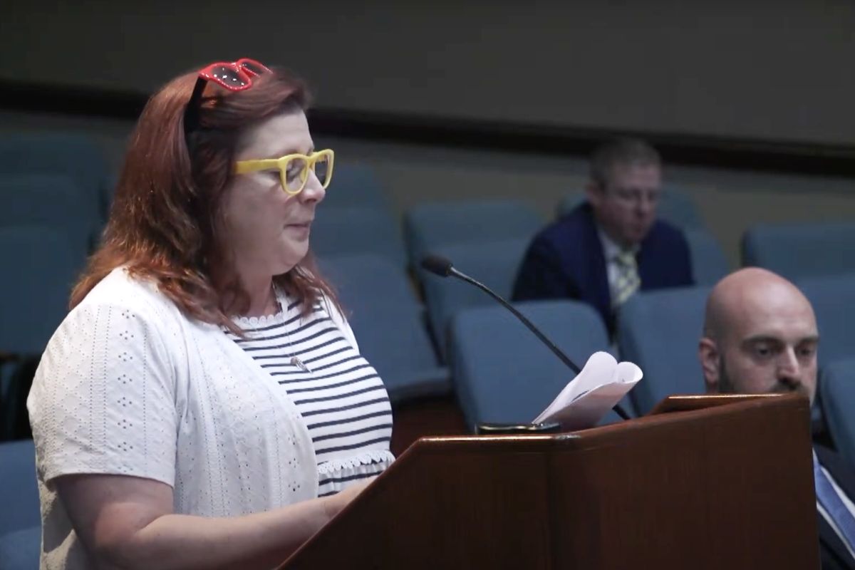 Abby Means stands at a podium holding a packet of papers that have been stapled together. Means is a White woman with shoulder length red hair.