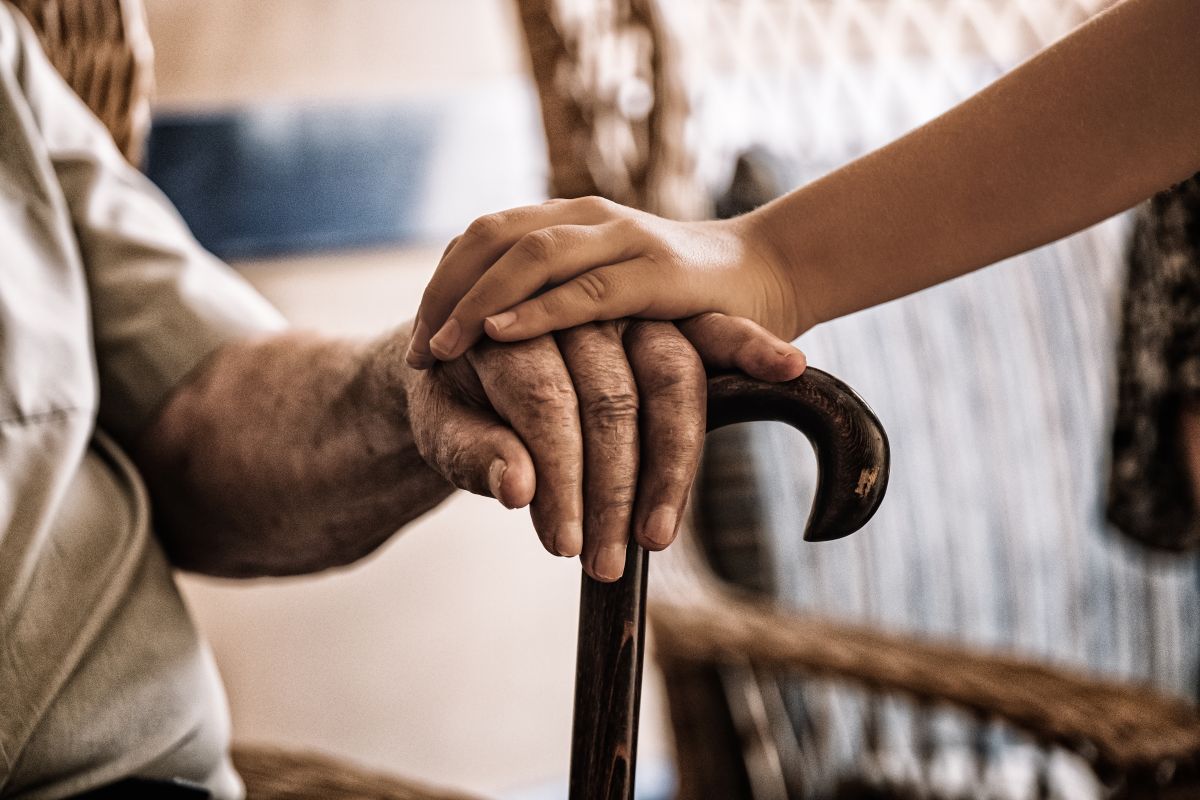 An elderly person rests their hand on a cane. Another person rests their hand on top. 