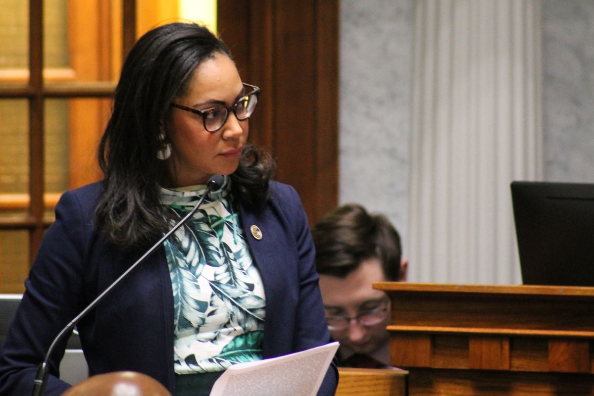 Senator Andrea Hunley stands behind a podium and speaks into a microphone while holding a stack of papers. She is a Black woman with dark hair and glasses, wearing a suit.