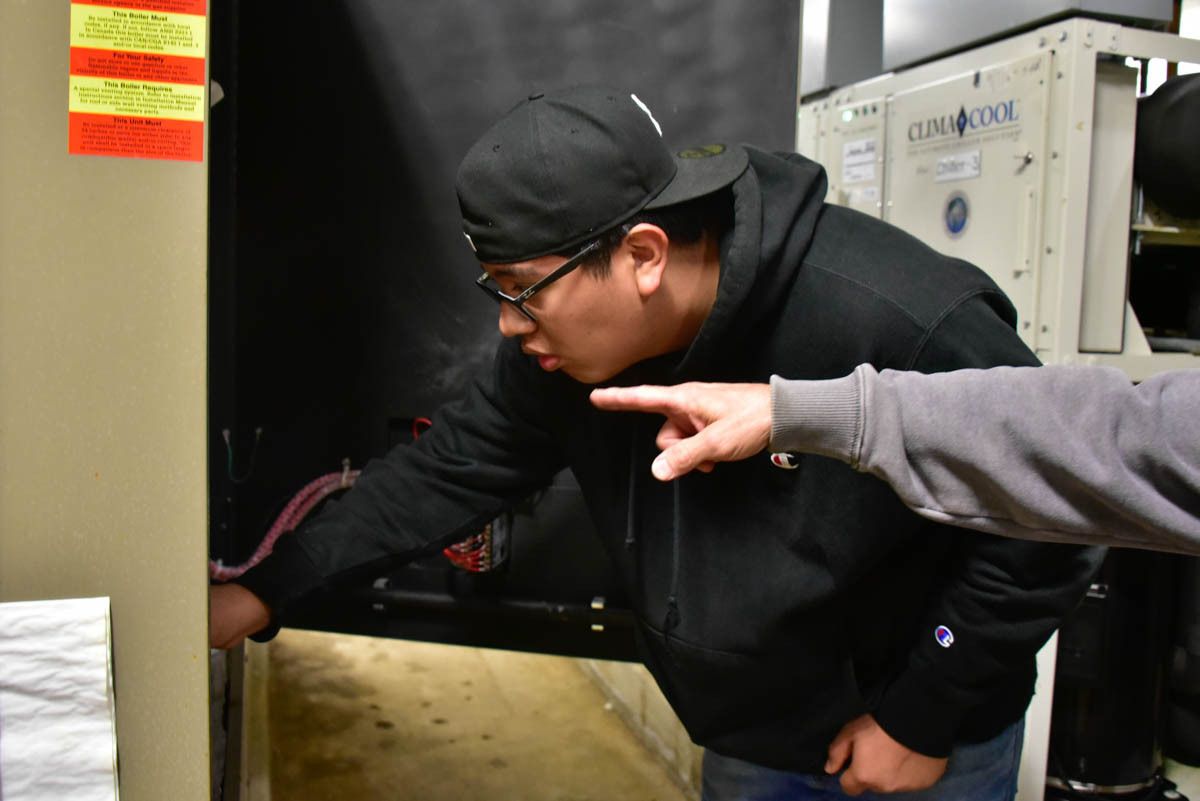 A young man is wearing a black hoodie and black baseball cap as he works on an HVAC unit under the supervision of another person, whose hand and sleeve are the only things visible.