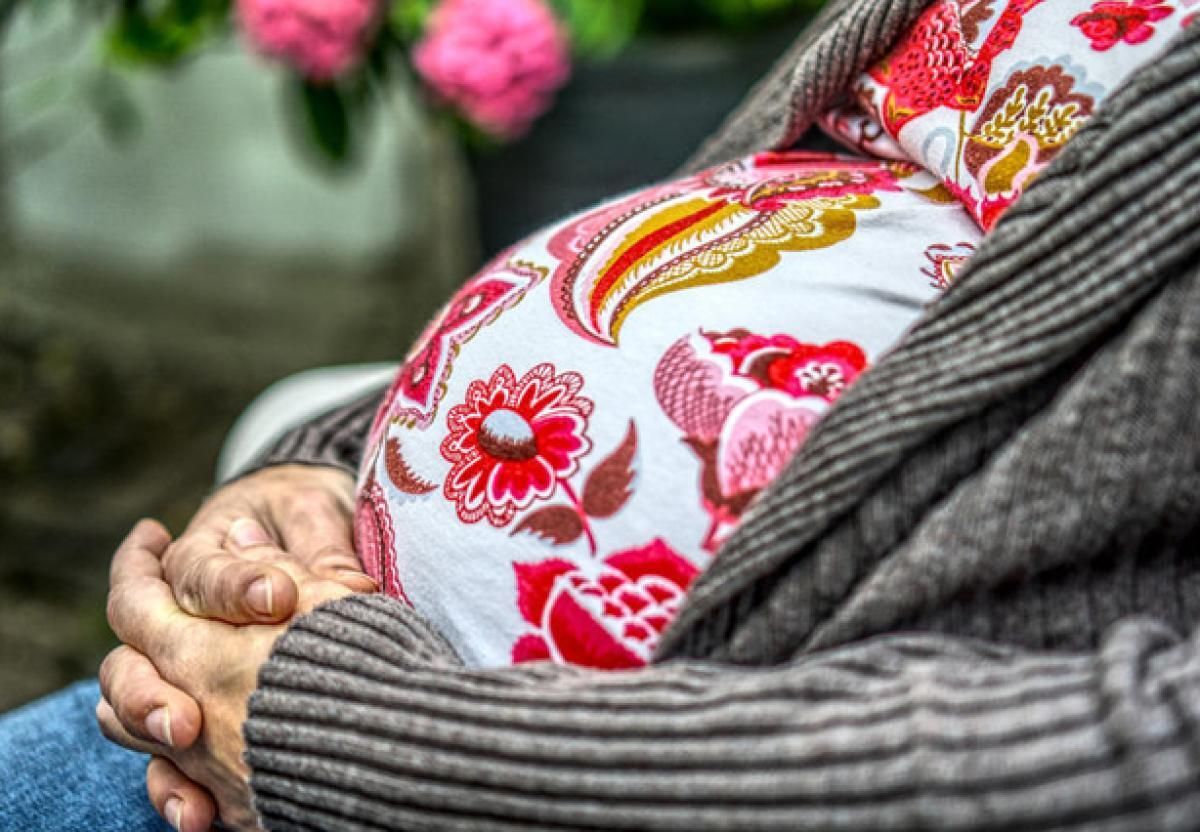 A pregnant person wears a shirt with a white base with pink and red flowers all across the front of the shirt. They wear a gray sweater layered on top of the shirt with their hands folded. 