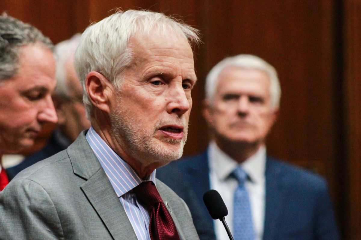 Representative Bob Behning speaks at a microphone. He is a White man with white hair and stubble. He wears a suit with a dark red tie.