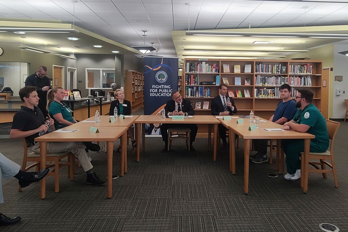 People sit around tables in Ivy Tech Valparaiso's library. Secretary Cardona is in the middle, listening to a student in scrubs speak.