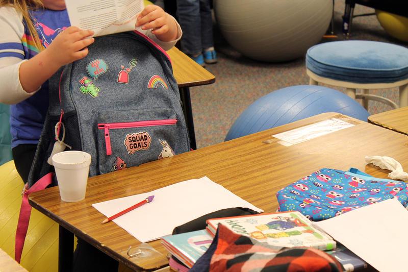 A student puts a piece of paper into her backpack while sitting at a table in a school.