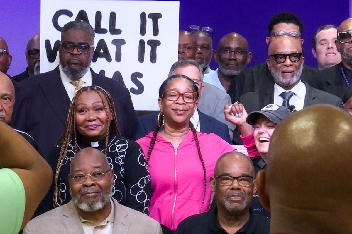 Attendees of the gathering pose for a group photo. A Black woman in the foreground holds her fist up. Behind her and several others is a sign that says "Call it what it was".