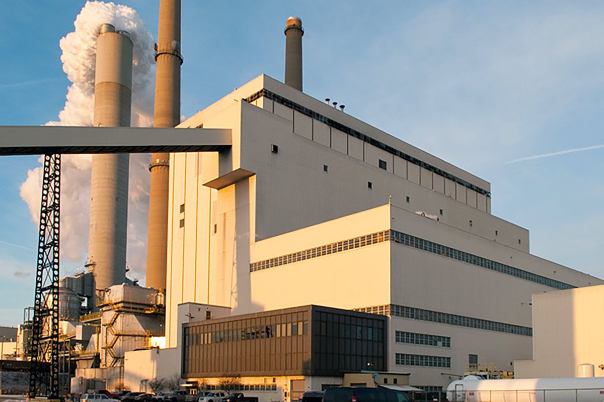 The Cayuga Generating Station in Vermillion County. A large, modular white building is wrapped with grey lines. A long, covered belt feeds into the top of the building. Behind it, stacks billow smoke.