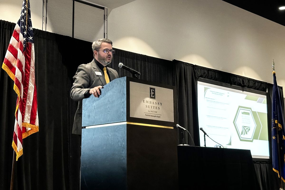 Dan Hardesty speaks into a microphone on a podium. He wears a suit with a yellow tie and glasses and has a beard.