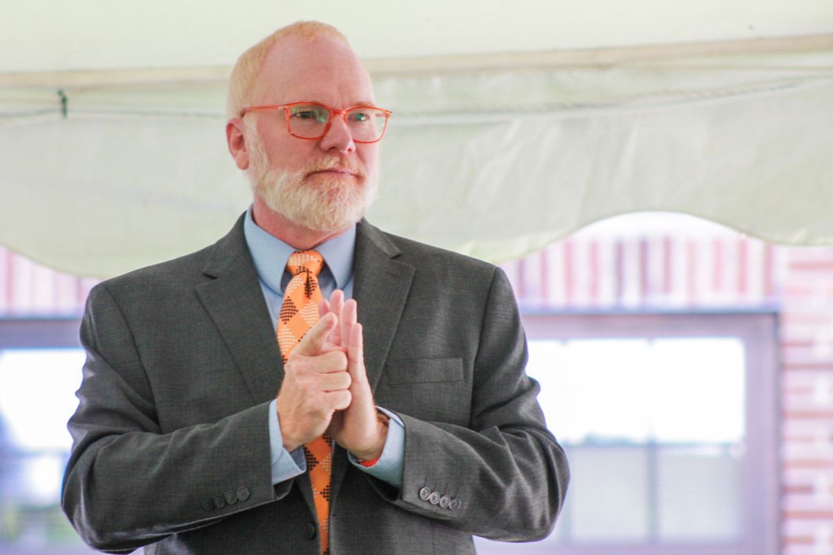 David Geeslin signs while making a speech. Geeslin is a White man with red and white hair and beard. He is wearing glasses and a gray suit, blue shirt and orange checkered tie