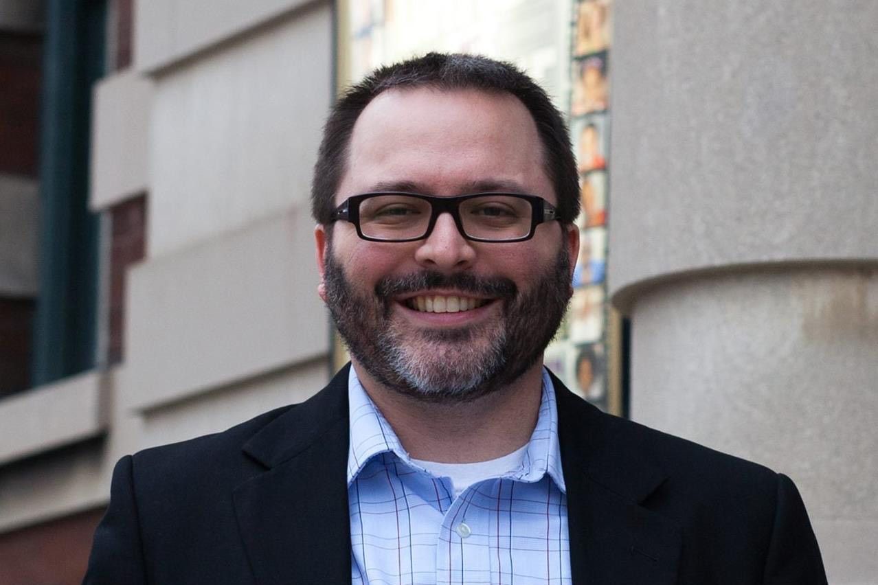 Derek Camp is a White man with dark hair and beard. He is wearing glasses and a black blazer over a light blue shirt.