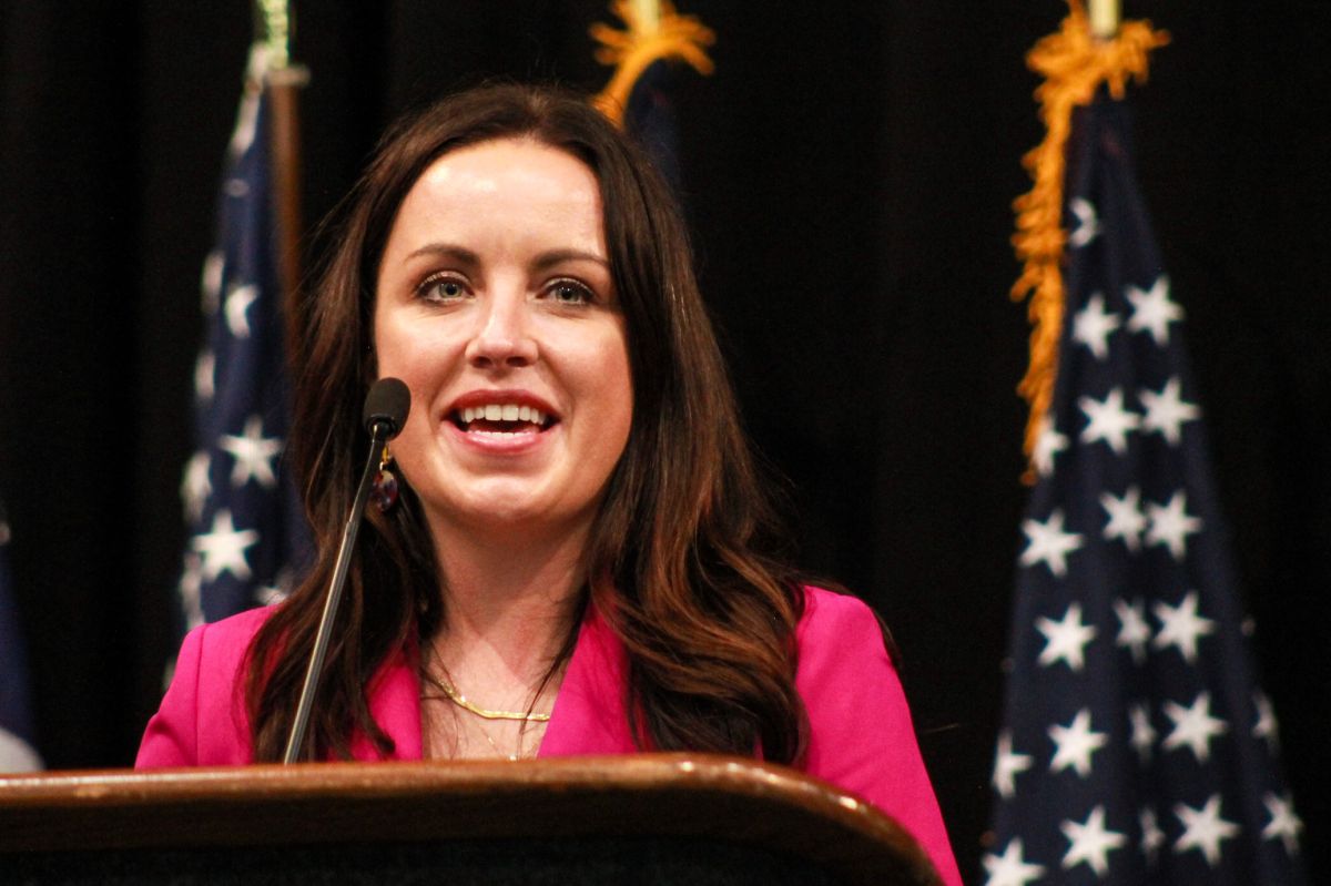 Destiny Wells speaks into a microphone at a lectern. There are American flags standing behind her. Wells is a White woman with dark brown hair.