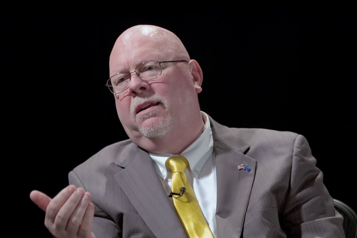 A screenshot from a video interview with Donald Rainwater. Rainwater is a White man, bald with a white goatee. He is wearing glasses and a tan suit with a white shirt and yellow tie.